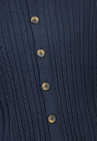 Granatowy, dziergany kardigan z wzorem w warkocze, sześcioma brązowymi guzikami z przodu oraz prążkowaną fakturą na całej powierzchni.