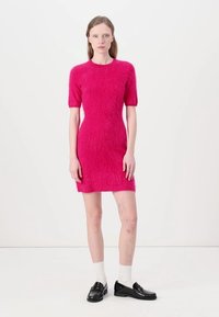 Fuzzy rosa Kleid mit kurzen Ärmeln, taillierter Silhouette und gerippter Textur. Kombiniert mit schwarzen Schuhen und weißen Socken steht das Model vor einem weißen Hintergrund.
