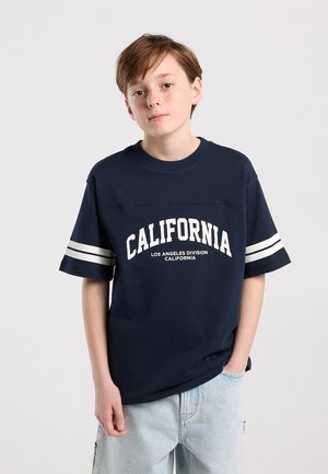 Jeune personne aux cheveux bruns courts portant un t-shirt bleu marine « California » et un jean bleu clair, debout devant un fond uni.