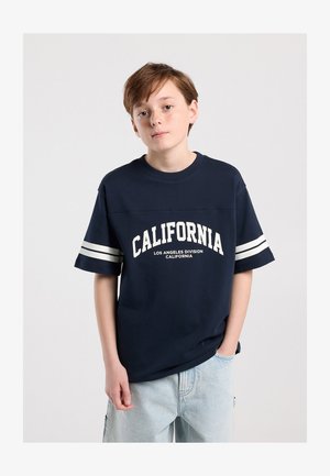 Persona joven con cabello castaño corto, vistiendo una camiseta azul marino con la palabra "California" y vaqueros azul claro, de pie frente a un fondo liso.