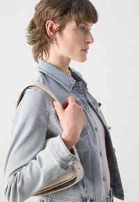Jeans jakna svijetlo plavog pranja, s dugmetima sprijeda i na manžetama. Ruka drži kremastu torbu za rame koja ističe svoju glatku teksturu.