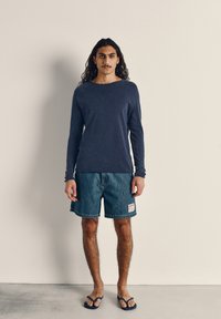 Tamsiai mėlynas medvilninis ilgomis rankovėmis marškinėlis, džinsiniai šortai su etikete ir mėlyni šlepetės, pavaizduoti ant asmens neutralioje fone.