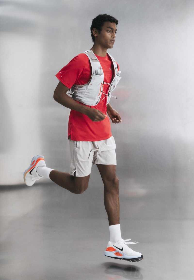Läufer mit rotem Shirt, weißen Shorts, weißen Sneakers und einer hellgrauen Trinkweste joggt drinnen vor schlichtem grauem Hintergrund.
