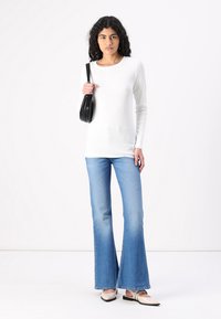 Top bianco a maniche lunghe, jeans svasati azzurri chiari, borsa nera e scarpe beige slip-on. Texture lisce e design aderente.