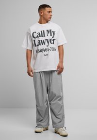 Hvit oversized t-skjorte med svart tekst "Ring min advokat 1(888)464-7065" kombinert med løse grå cargo-bukser og kremfargede sneakers.