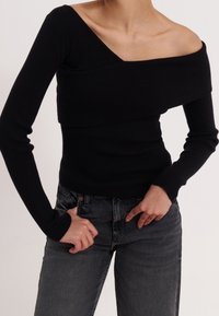 Person trägt einen schwarzen, schulterfreien, gerippten Pullover und eine dunkelgraue Jeans, während er mit den Händen die Jeans an der Taille anpasst.