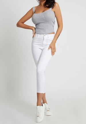 Grauer gerippter Crop-Top mit breiten Trägern, kombiniert mit weißen, mittelhoch geschnittenen Skinny-Jeans. Das Model trägt weiße Sneakers und steht vor einem neutralen Hintergrund.