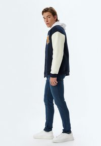 Chaqueta varsity de la marina con mangas crema, botones en el frente y parche de letra naranja. Lleva puesta sobre una sudadera blanca, combinada con jeans azules y zapatillas blancas.