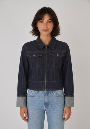 Donna con capelli scuri fino alle spalle indossa una giacca di jeans scura con chiusura a zip e polsini piegati e jeans azzurri, in piedi davanti a uno sfondo semplice.