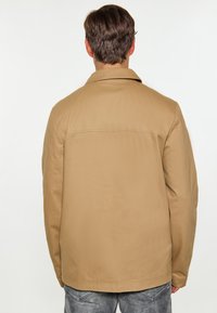 Tan jakke med en struktureret krave, lige snit og glat tekstur, parret med lysegrå jeans. Synlig syning på jakken.