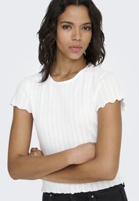 Witte ribgebreide katoenen T-shirt met geschulpte mouwen en zoom, met een gestructureerd patroon. Model poseert met gekruiste armen, draagt een zwarte broek.