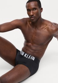 Črne bombažne spodnjice z belim pasom z logotipom "Calvin Klein", ki se ponašajo z gladko teksturo in prilegajočim dizajnom. Model sedi in prikazuje izdelek.