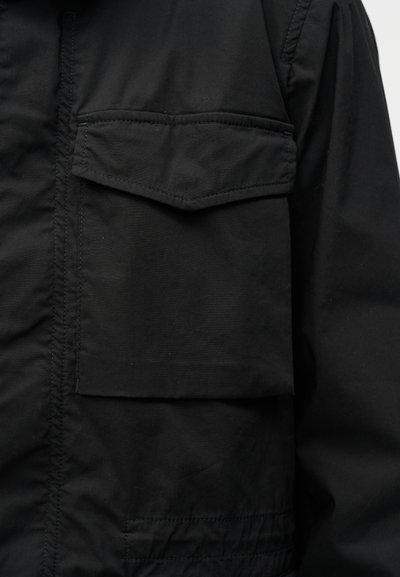 Μαύρο μπουφάν σε κοντινό πλάνο που δείχνει μπροστινή τσέπη με καπάκι και εμφανείς ραφές σε ελαφρώς τσαλακωμένο ύφασμα.