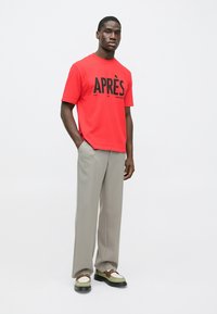 Sarkans īss piedurkņu T-krekls ar lielu melnu uzrakstu “APRÈS”, kopā ar gaiši pelēkām plīsētām biksēm un daudzkrāsainām sporta kurpēm.