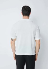 Polo shirt blanc à manches courtes avec motif texturé, col classique et coupe décontractée, associé à un pantalon noir.