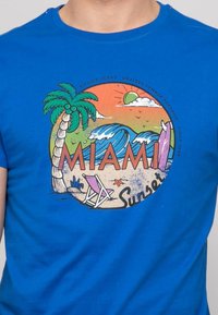 T-shirt en coton bleu avec un graphique circulaire représentant des palmiers, des vagues et une scène de plage. Texte "MIAMI Sunset" en orange vif.
