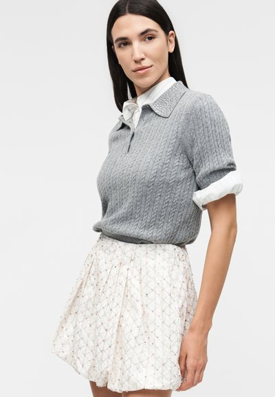 Γυναίκα με ίσια σκούρα μαλλιά φοράει γκρι πλεκτό πουλόβερ με κοντά μανίκια πάνω από ένα λευκό πουκάμισο, συνδυασμένο με λευκή φούστα με σχέδια.