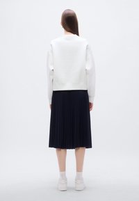 Sweat-shirt en coton blanc avec poignets et ourlet côtelés, associé à une jupe plissée bleu marine arrivant au genou. Le modèle porte des baskets blanches et des chaussettes.