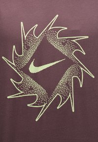 Το λογότυπο της Nike στο κέντρο ενός διαμαντιού με ακίδες, με διακεκομμένα και υφασμένα μοτίβα σε φόντο από μπορντό ύφασμα.