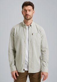 Camisa de botón a rayas en verde claro y blanco, con un bolsillo en el pecho, mangas largas y dobladillo redondeado. Llevada sobre una camiseta blanca.