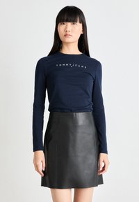Tengerészkék, hosszú ujjú felső "TOMMYJEANS" logóval, fekete bőr mini szoknyával párosítva, amely sima textúrájú és diszkrét varrásokkal rendelkezik.