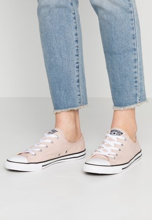 Pieds portant des baskets Converse All Star basses rose clair avec des lacets blancs et un jean en denim aux ourlets effilochés.