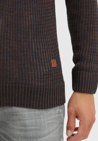 Strikpullover i mørkeblå og brune nuancer; har ribbet struktur, rund hals og logo-patch på sømmen.