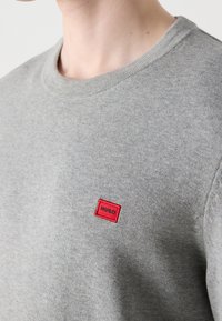 Personne portant un pull ras du cou gris clair avec un petit patch rectangulaire rouge portant l'inscription "HUGO" sur la poitrine.
