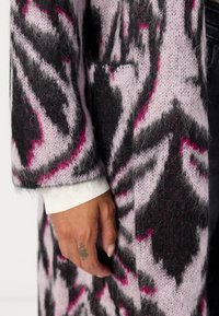 Knitted coat in shades of black, pink, and lavender, featuring a textured pattern. The sleeve ends with a white cuff, showcasing a relaxed silhouette.