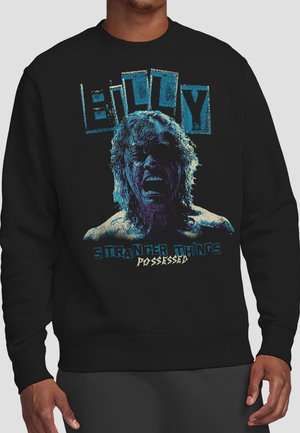 Man wearing black sweatshirt with graphic of distressed face, text "Elliot," and "Stranger Things Possessed" in blue and cream colors.