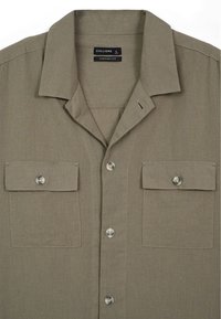 Chemise à manches courtes olive avec un col rabattable, deux poches avant et des boutons transparents. Fabriquée en tissu léger.