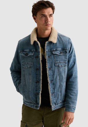 Jeune homme portant une veste en jean bleue avec une doublure polaire beige et un pantalon cargo vert, regardant légèrement vers la droite, les mains dans les poches.