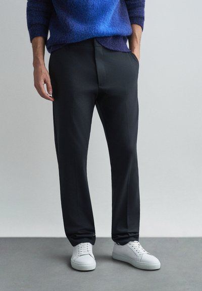 Calças pretas ajustadas com uma textura suave, apresentando bolsos laterais, combinadas com ténis brancos e um suéter de malha azul.