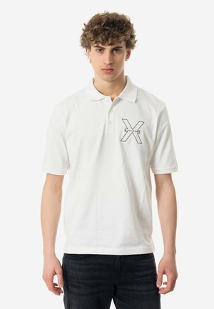 Camisa polo de algodón blanca con collar abotonado. Presenta un logotipo gráfico negro en forma de 'X' en el lado izquierdo y mangas cortas.