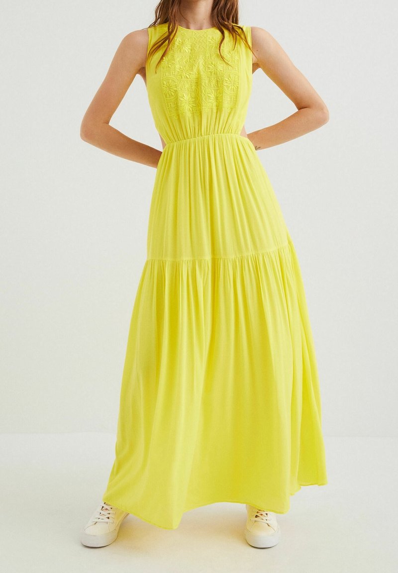 Femme portant une robe maxi jaune sans manches avec un corsage brodé et des baskets blanches, debout les mains sur les hanches.