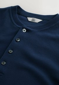 Top din tricot ribbed de culoare albastru marin, cu guler rotund și patru nasturi negri pe față, eticheta vizibilă în interiorul gulerului.