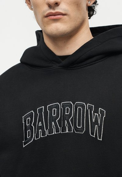 Νέος άνδρας που φοράει μαύρο φούτερ με κουκούλα και λευκά επισημασμένα γράμματα πάνω στο στήθος που σχηματίζουν τη λέξη «BARROW», κοιτάζοντας μπροστά με ουδέτερη έκφραση.