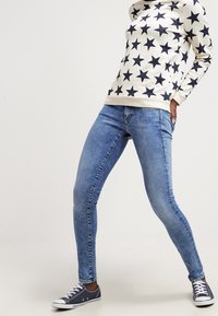 Suéter beige con patrón de estrellas en azul marino, combinado con jeans ajustados azules y zapatillas en azul marino. Conjunto informal con colores simples y contrastantes.