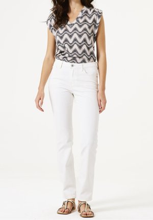 Vrouw met een zwart-wit patroonloze mouwloze top, witte high-waisted broek en beige platte sandalen met bandjes, staand op een witte achtergrond.