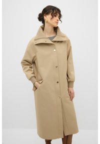 Beige knielang jasje met een hoge kraag, knoopsluiting en zijzakken. Voorzien van elastische manchetten en hardware-details in goudkleur.