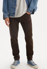 Pantaloni in corduroy marroni con vestibilità slim, abbinati a una maglietta grigia e a una giacca in denim blu, completati da sneakers blu.