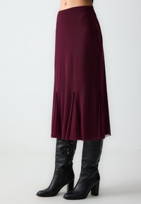 Jupe midi bordeaux avec un design plissé, en tissu léger, associée à des bottes en cuir noires à talons montantes.