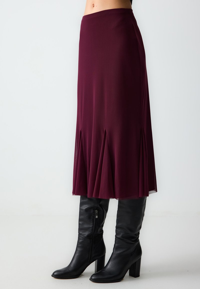 Gonna midi bordeaux con design a pieghe, realizzata in tessuto leggero, abbinata a stivali neri in pelle con tacco al ginocchio.