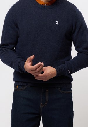 Mand iført navyblå sweater med hvid polo-spiller logo over orange skjorte med krave og mørkeblå jeans, hænderne foldet foran.