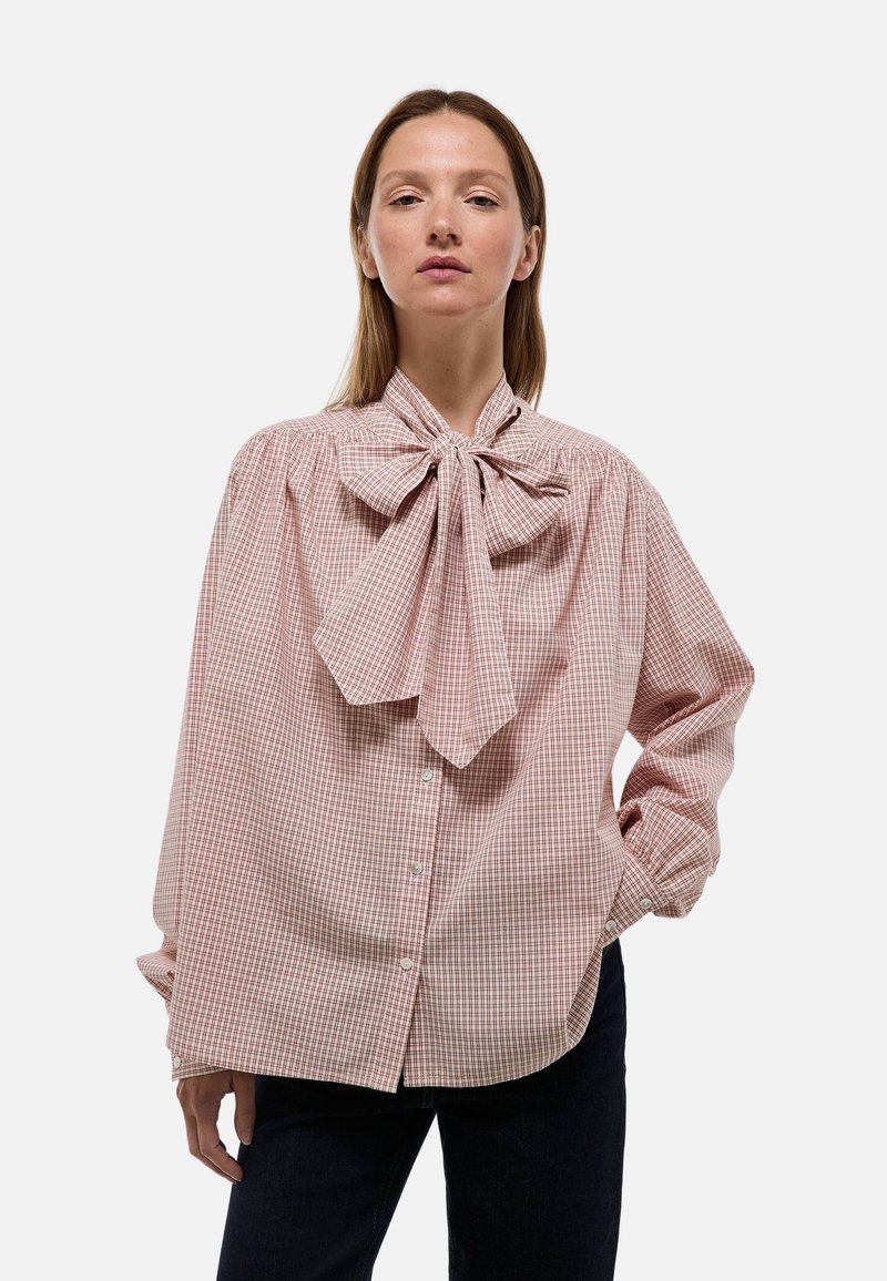 Blouse à carreaux roses et blancs avec un grand col en nœud, manches longues et fermeture par bouton. Le tissu semble léger et texturé.