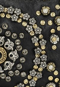 Zwart textured materiaal met decoratieve elementen, waaronder zilveren en gouden bloemen, metalen studsen en ronde kristallen, gerangschikt in een patroon.