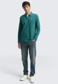 Langärmliges türkisfarbenes Polohemd mit einem weißen Kragen, versehen mit einem kleinen Logo, kombiniert mit hellblauen Jeans und weißen Sneakern.