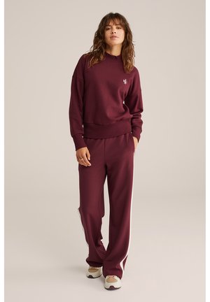Felpa bordeaux con bordo a costine, abbinata a pantaloni della tuta bordeaux con strisce laterali bianche e scarpe sportive beige.