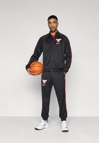 Μαύρη αθλητική φόρμα με κόκκινες λεπτομέρειες, με λογότυπο στο μπουφάν και το παντελόνι. Ο παίκτης κρατάει μπάλα μπάσκετ φορώντας λευκά αθλητικά παπούτσια.