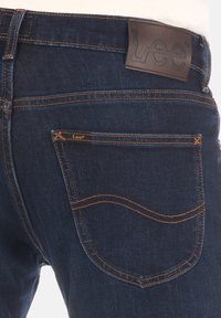 Donkerblauwe denim jeans met een leren label met de naam "Lee". Oranje stiksels omlijnen de achterzak en naden. Klassiek, slim fit ontwerp.